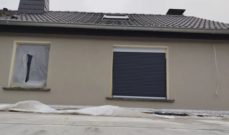 Exterior of a residential building with a beige facade, featuring a small window with cover on the left and a larger window with dark shutters on the right, with metal roofing above.