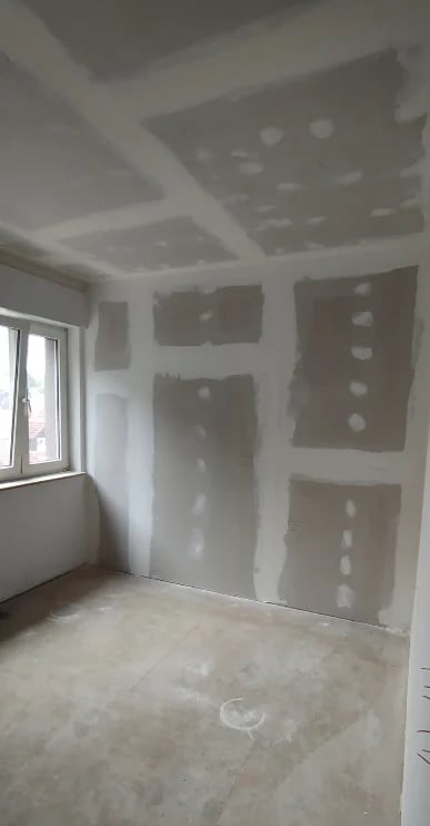 Empty room under construction with drywall installation, window on left wall, and unfinished ceiling