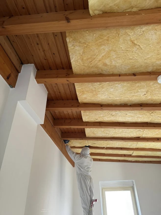 Interior ceiling with exposed wooden beams and yellow insulation batts, white drywall walls, and plastic sheeting on column