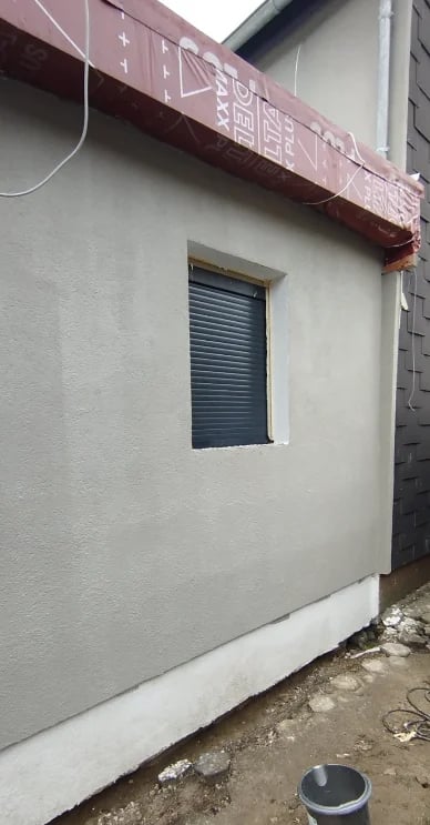 Small white building exterior with closed blue metal shutter window and faded red awning overhead