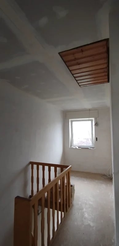 Interior of unfinished room with wooden staircase railing, white walls, window, and recessed ceiling light.