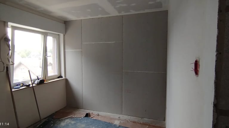 Construction site interior with drywall panels mounted on walls, window with plastic sheeting, and protective floor covering.